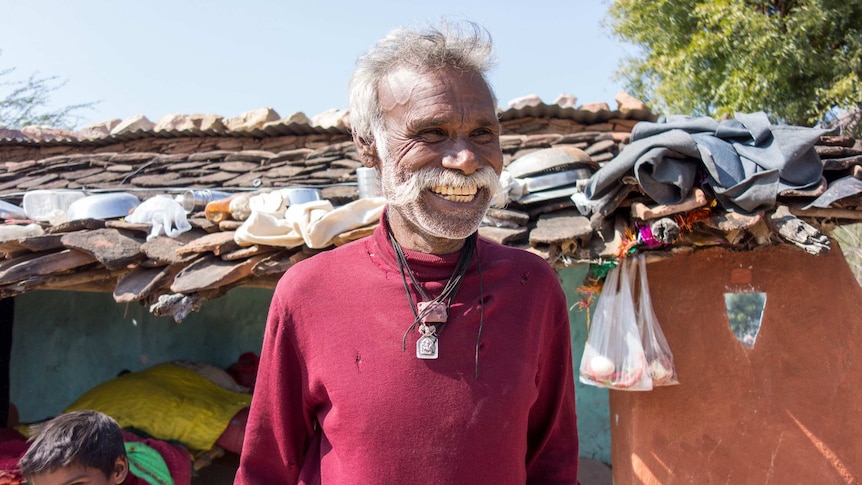 A man with a large white moustache and a red top smiles with a stone house in the background.