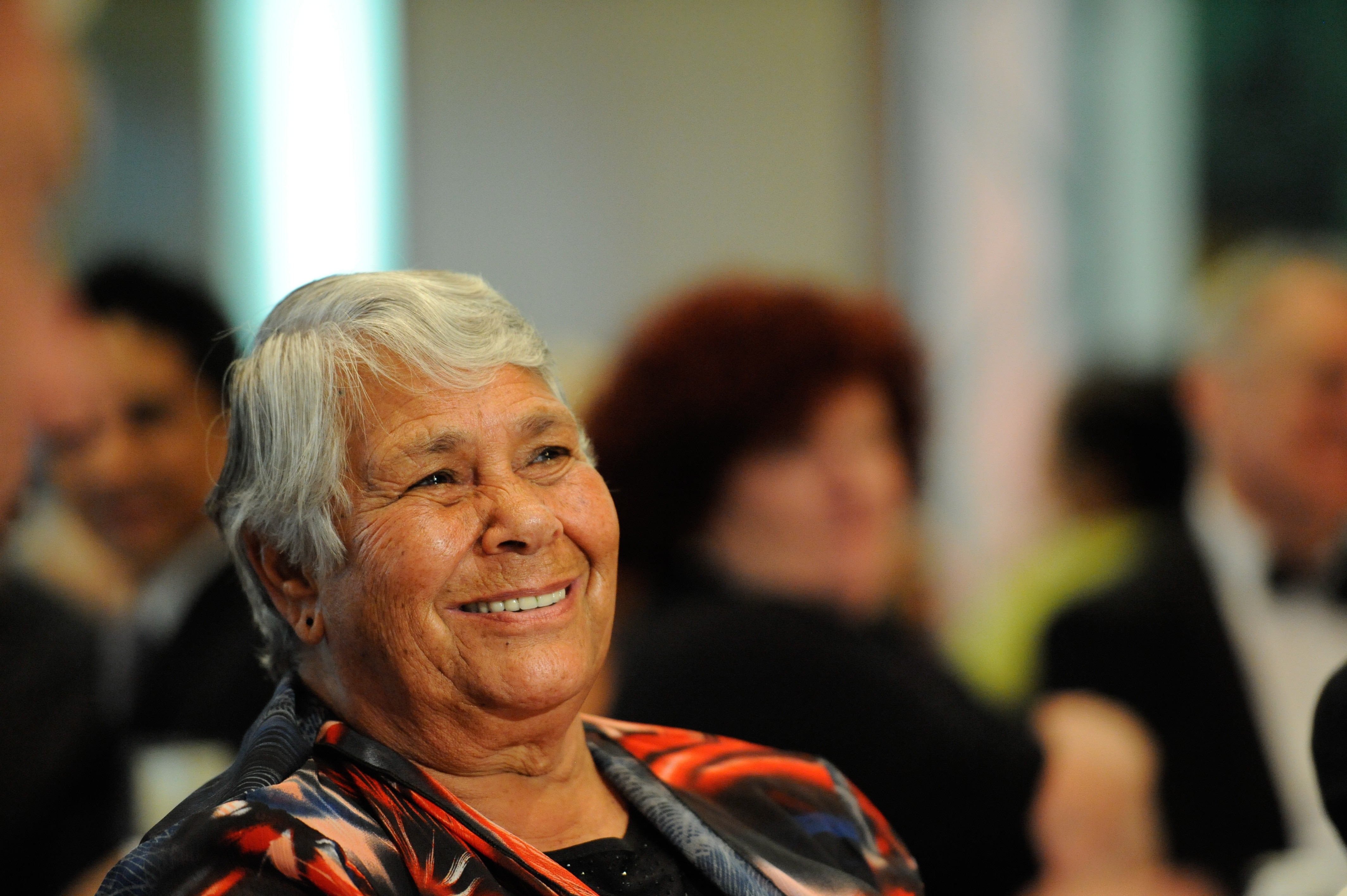 A close up of an Indigenous Australian woman sitting and smiling indoors, with other people sitting behind her 