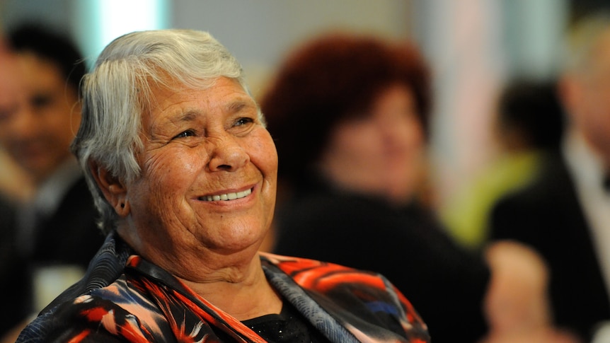 A close up of an Indigenous Australian woman sitting and smiling indoors, with other people sitting behind her 