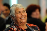 A close up of an Indigenous Australian woman sitting and smiling indoors, with other people sitting behind her 