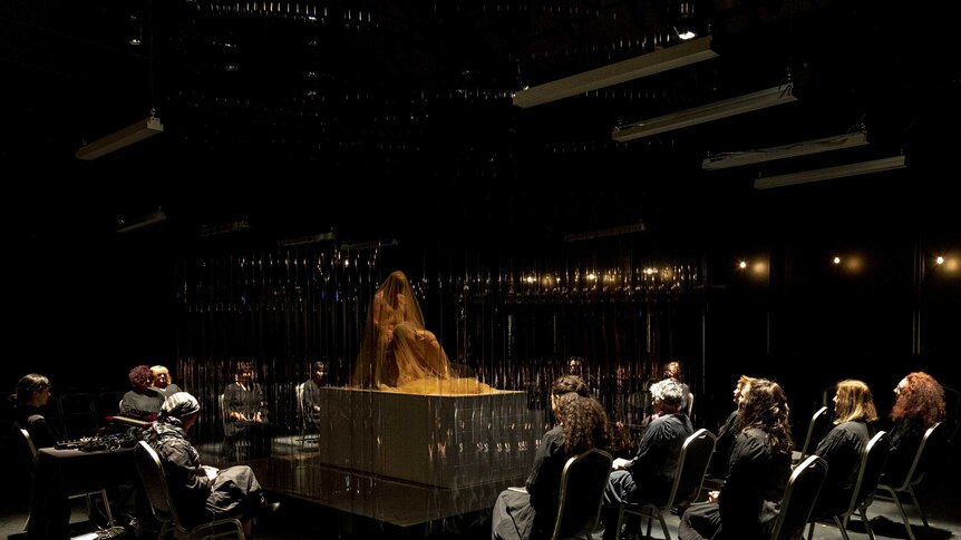 A group of women on chairs surround a stage with two shrouded figures