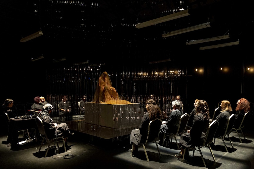 A group of women on chairs surround a stage with two shrouded figures