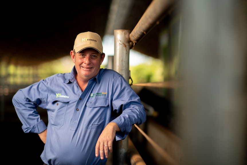 A man in a hat and a blue shirt leans against a cattle fence. 