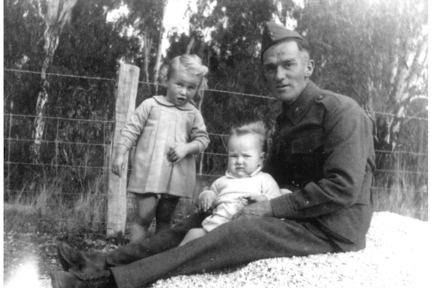 A black and white photo of a soldier holding a baby, while a toddler stands next to him in yard fenced with wire, trees.
