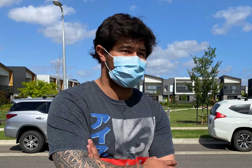 A man stands outside some townhouses on a sunny day wearing a mask