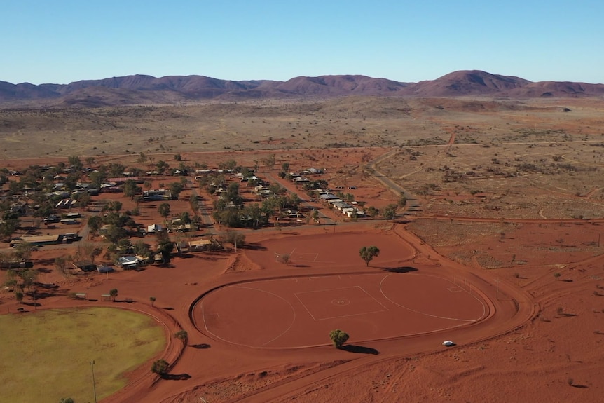 A red dirt football oval next to a grassed football oval and a small community with a mountain range in the background