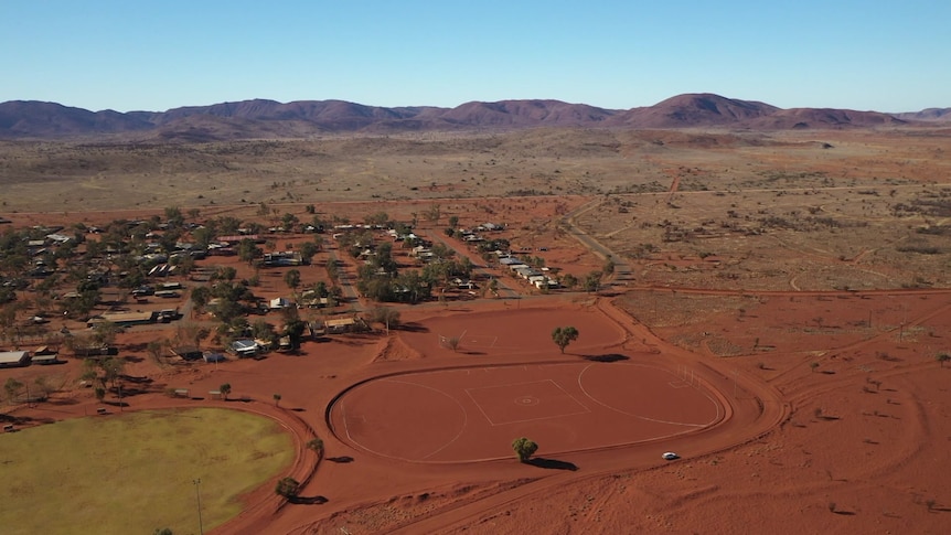 A red dirt football oval next to a grassed football oval and a small community with a mountain range in the background