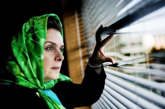 A woman in a head scarf peeks through the blinds.