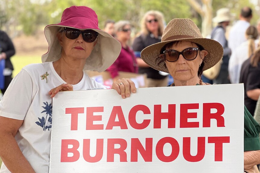 Two female teachers stand outside during a rally, posing for a photo with a sign reading 'teacher burnout' in red.