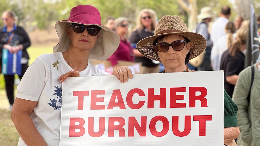 Two female teachers stand outside during a rally, posing for a photo with a sign reading 'teacher burnout' in red.