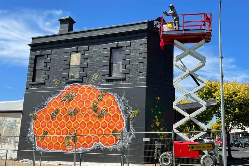A scissor lift being used to paint a mural of bees in honeycomb on a black building