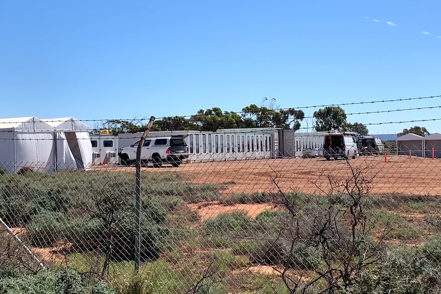 behind a barbed wire fence there are white tents and huts and a four wheel drive delivering something. shrubs in foreground.