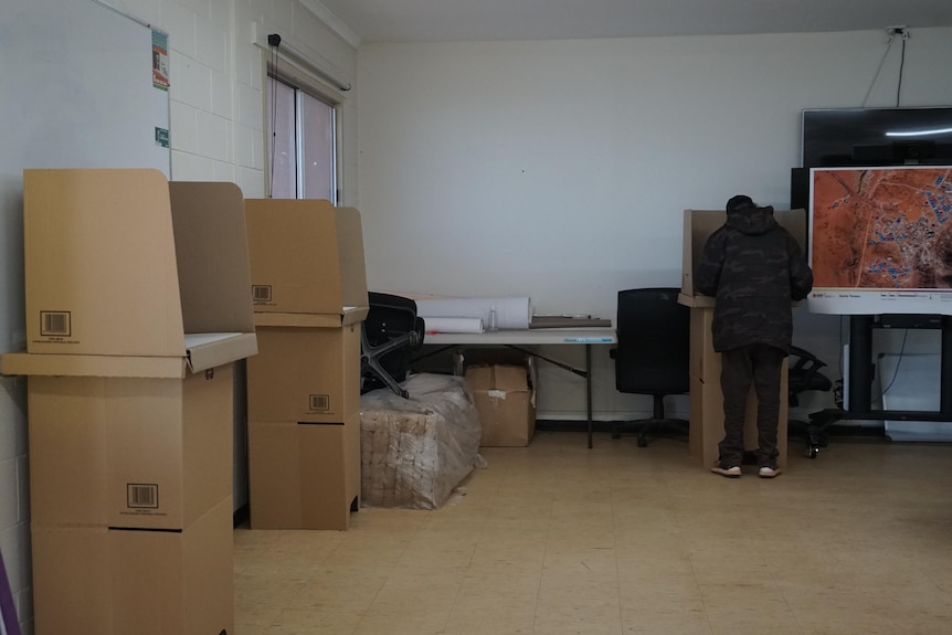 A picture of a room with three cardboard polling booths. A man is stood at one of the booths.