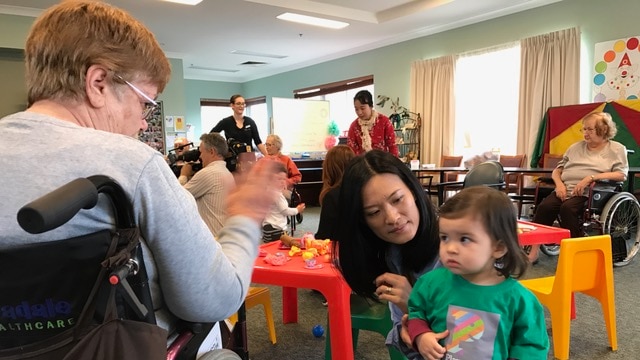 A mother encourages her young child to interact with an elderly aged care resident.