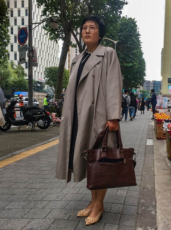 A North Korean woman with short black hair and round glasses holds a brown handbag and looks up into the distance on a sidewalk.
