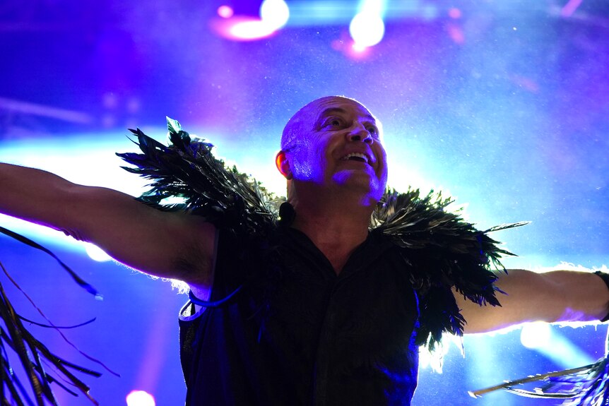 A bald man wearing a black singlet and feather neckpiece on a lit stage