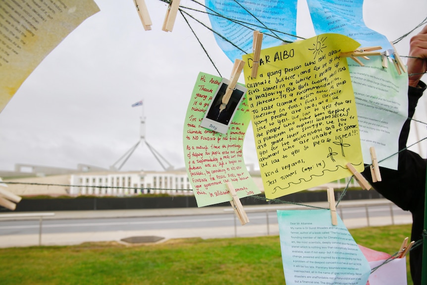 Няколко ръкописни бележки, ръчно закачени на тел, с сградата на парламента, която се вижда на заден план.
