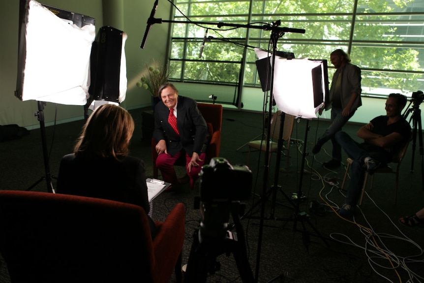 Humphries sitting chair surrounded by lights, camera and sound equipment with crew watching Hutcheon interviewing.