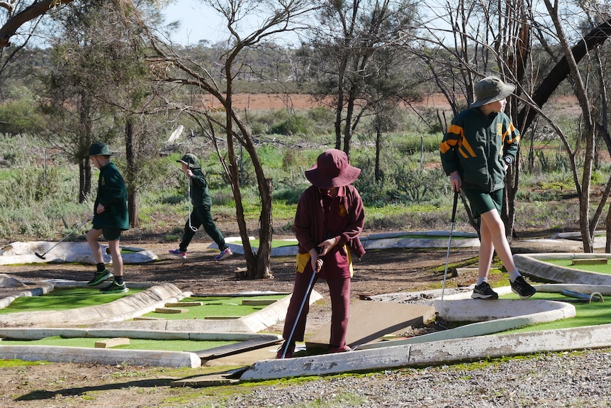 A boy in a maroon tracksuit and hat lines up a shot on a mini golf course with other students playing behind him.