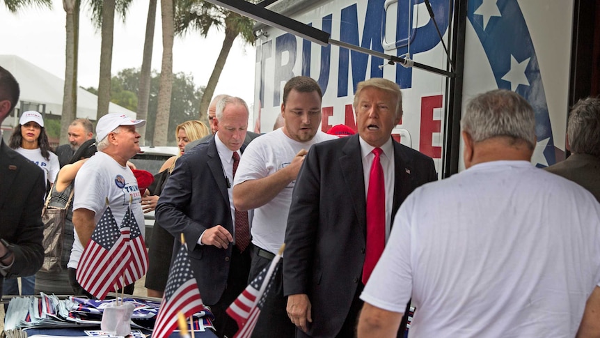 Donald Trump is standing outside an RV with half a dozen campaign staff or volunteers gather around him.