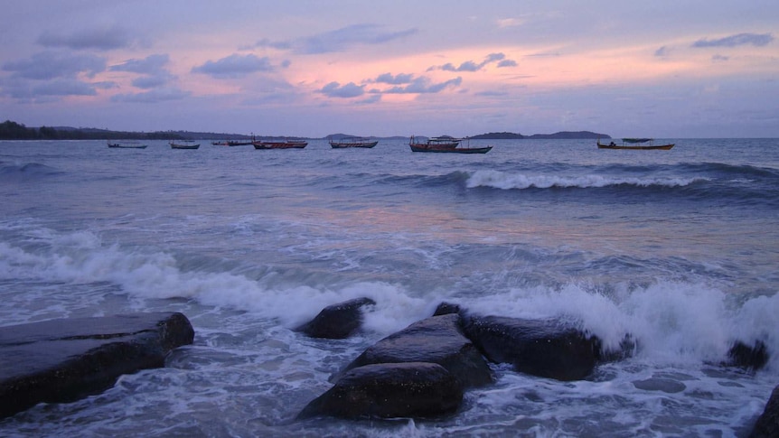Waves crashing on large rocks in shallow water at sunset. Fishing boats are anchored nearby, with mountains on the horizon.