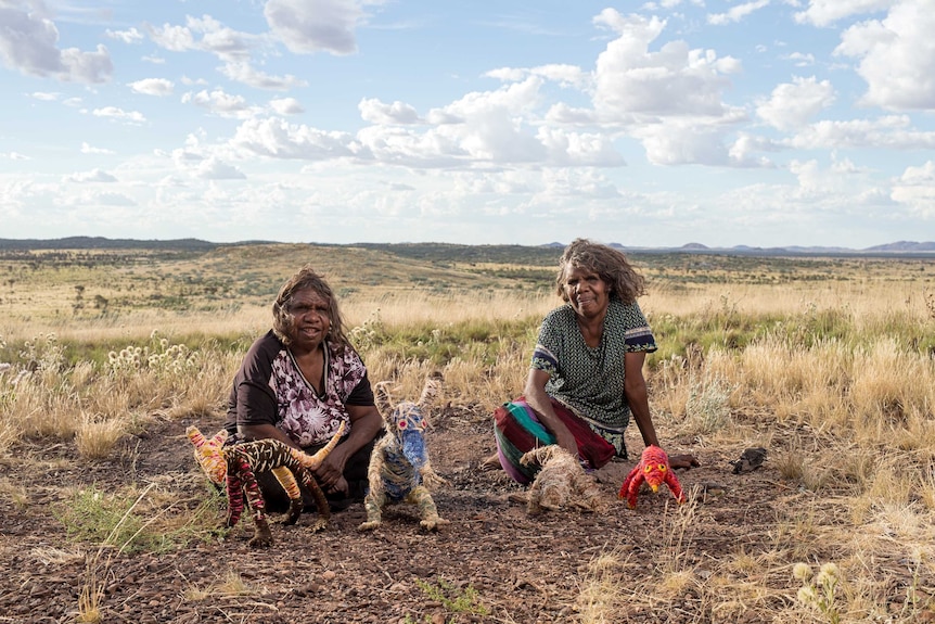Artists Roma Butler and Yangi Yangi Fox sitting in the Australian outback with sculptures made from native grasses