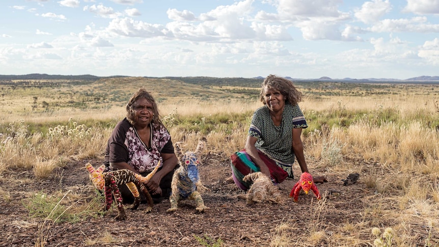 Artists Roma Butler and Yangi Yangi Fox sitting in the Australian outback with sculptures made from native grasses