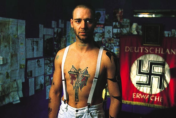 A bare-chested man with tattoos stands in front of a Nazi flag