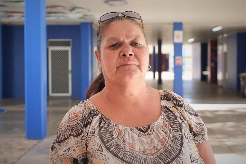 An unsmiling middle-aged Indigenous woman in a cream and black patterned blouse, stands in a large room with blue pillars.