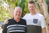 A tall young man has his arm around his father, both smiling in t-shirts, under trees