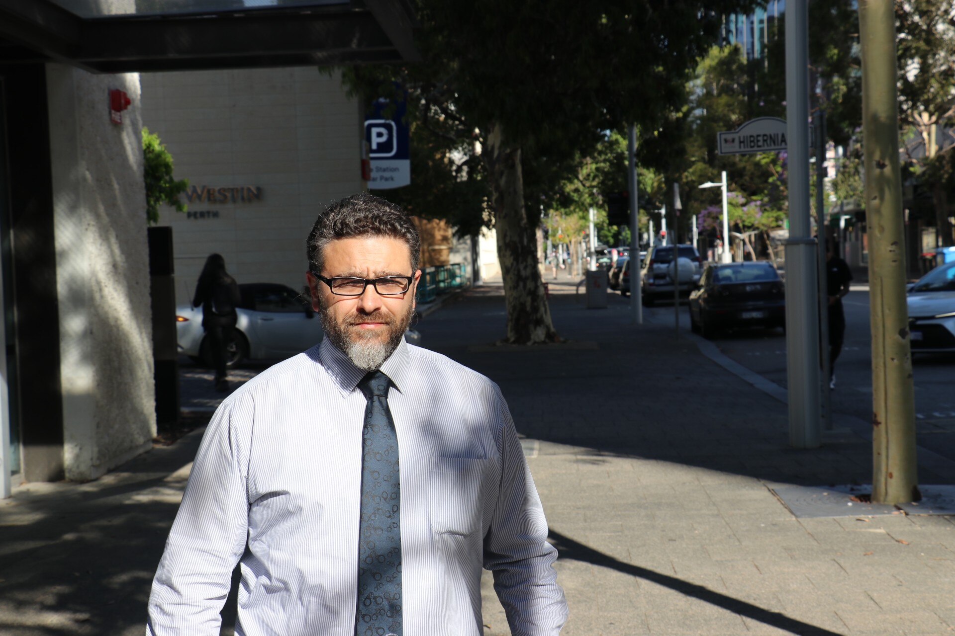 A man wearing a shirt, tie and glasses walks down a city street. 