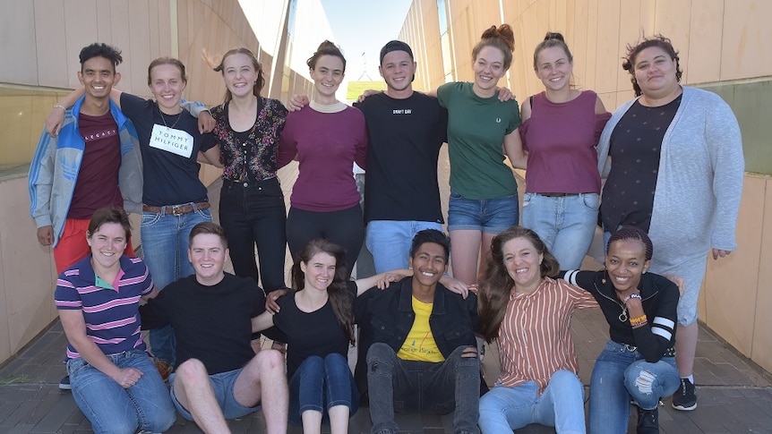 A group shot of 14 young adults smiling at the camera in front of a blurred corridor