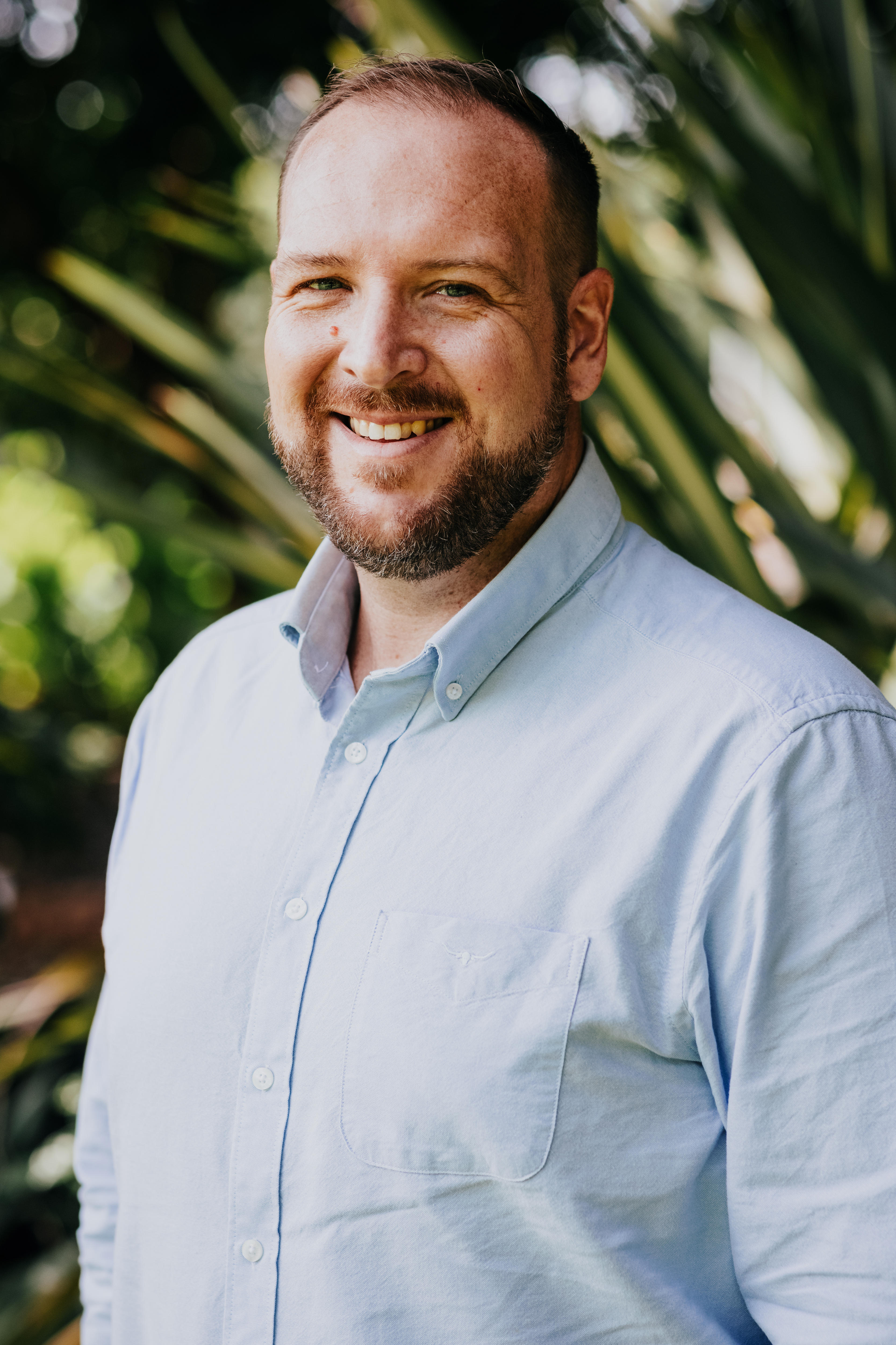 Justine Hales ceo stands in a light blue button up shirt smiling in front of some palm leaves