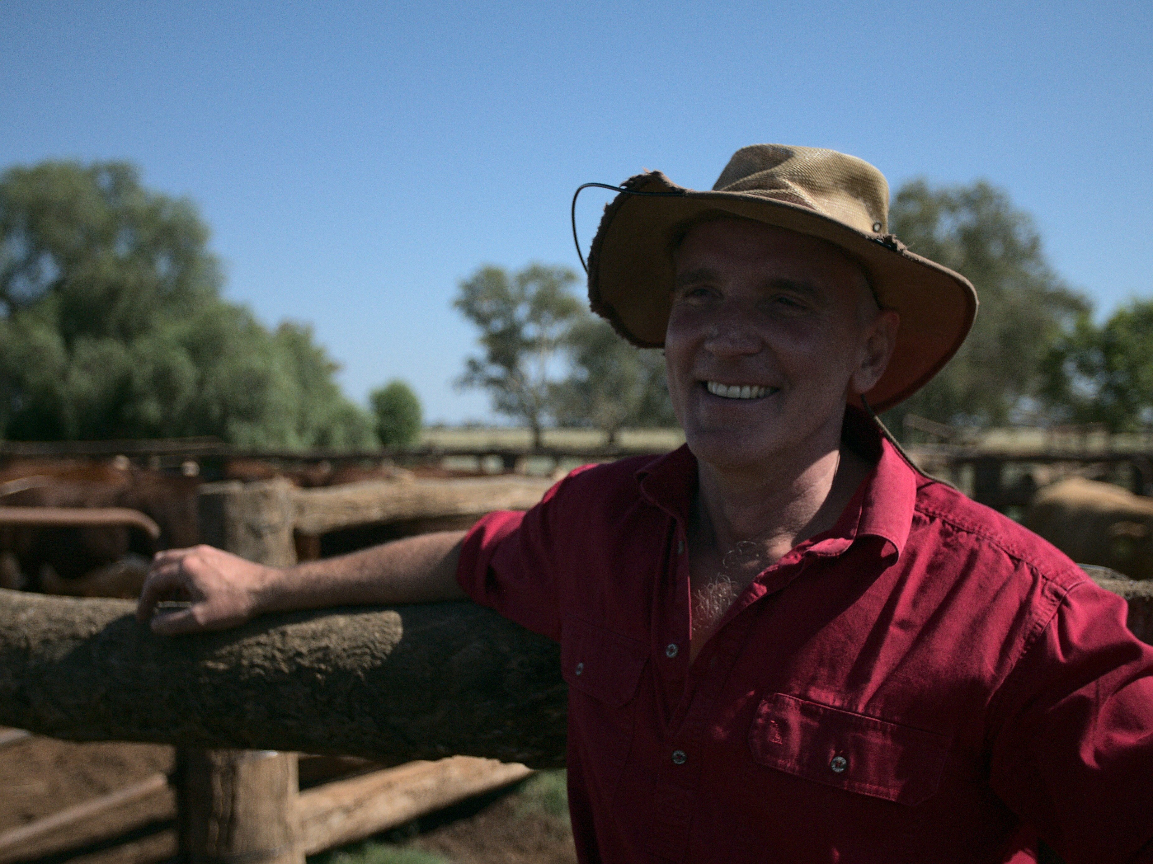 A man in a hat and red shirt smiles while leaning against the wooden fence of a cattle yard.