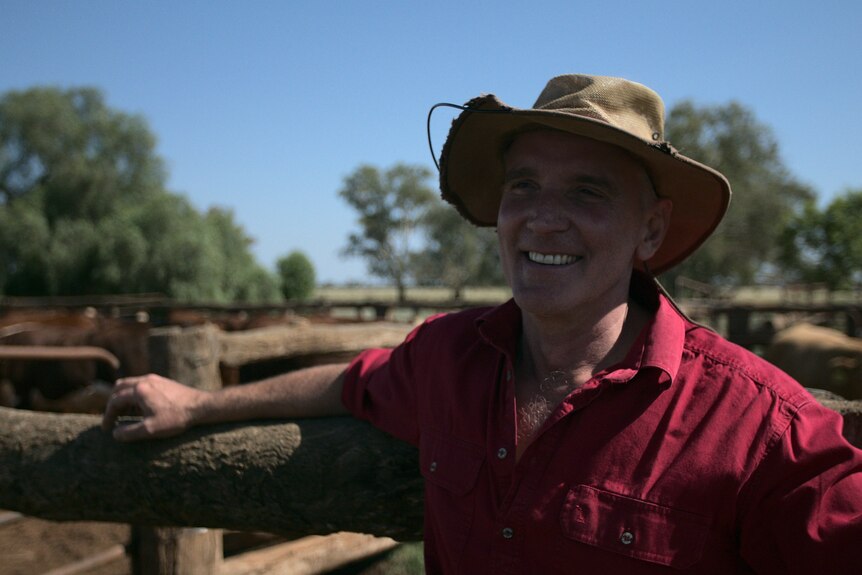 A man in a hat and red shirt smiles while leaning against the wooden fence of a cattle yard.