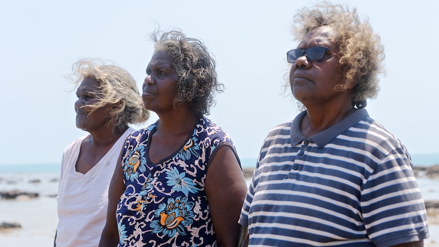 Three Indigenous women stand side by side looking into the distance with water behind them.