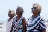 Three Indigenous women stand side by side looking into the distance with water behind them.