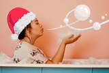 A woman sits in a bath wearing a Christmas hat and blowing bubbles.