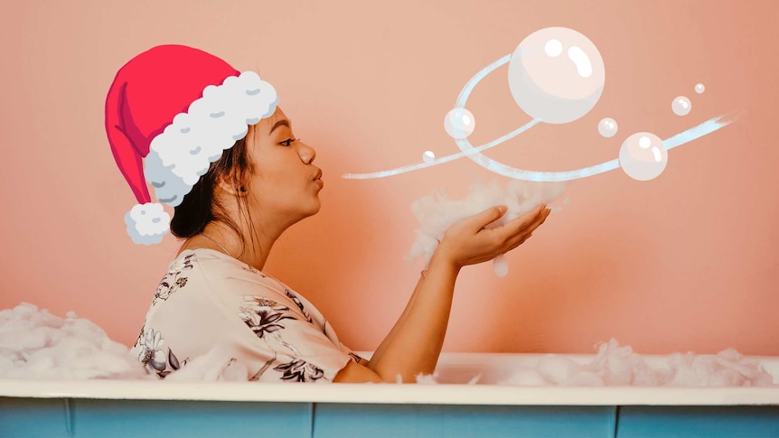 A woman sits in a bath wearing a Christmas hat and blowing bubbles.