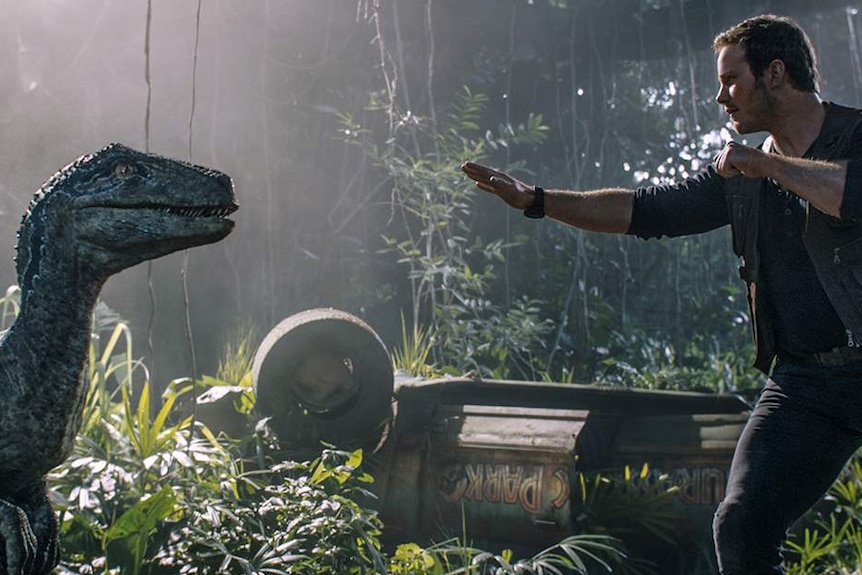 Chris Pratt's character holds a calming hand out to a human sized dinosaur standing on two legs.
