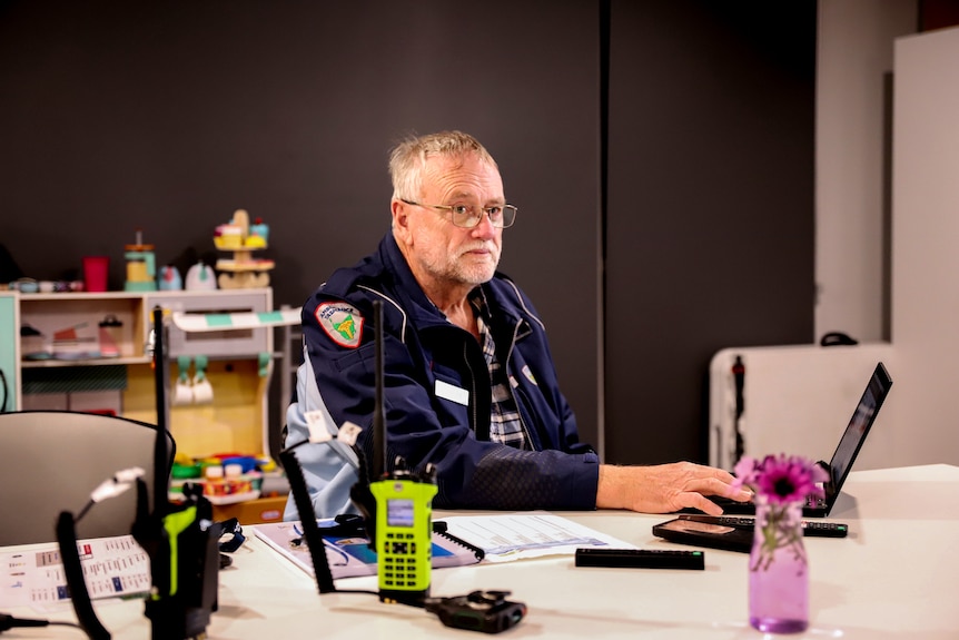 Older man with glasses sits at a table, looking across over laptop and walky talkies