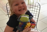 Izabella smiles into the camera while seated, she holds a green soft toy in a brightly coloured prosthetic hand.