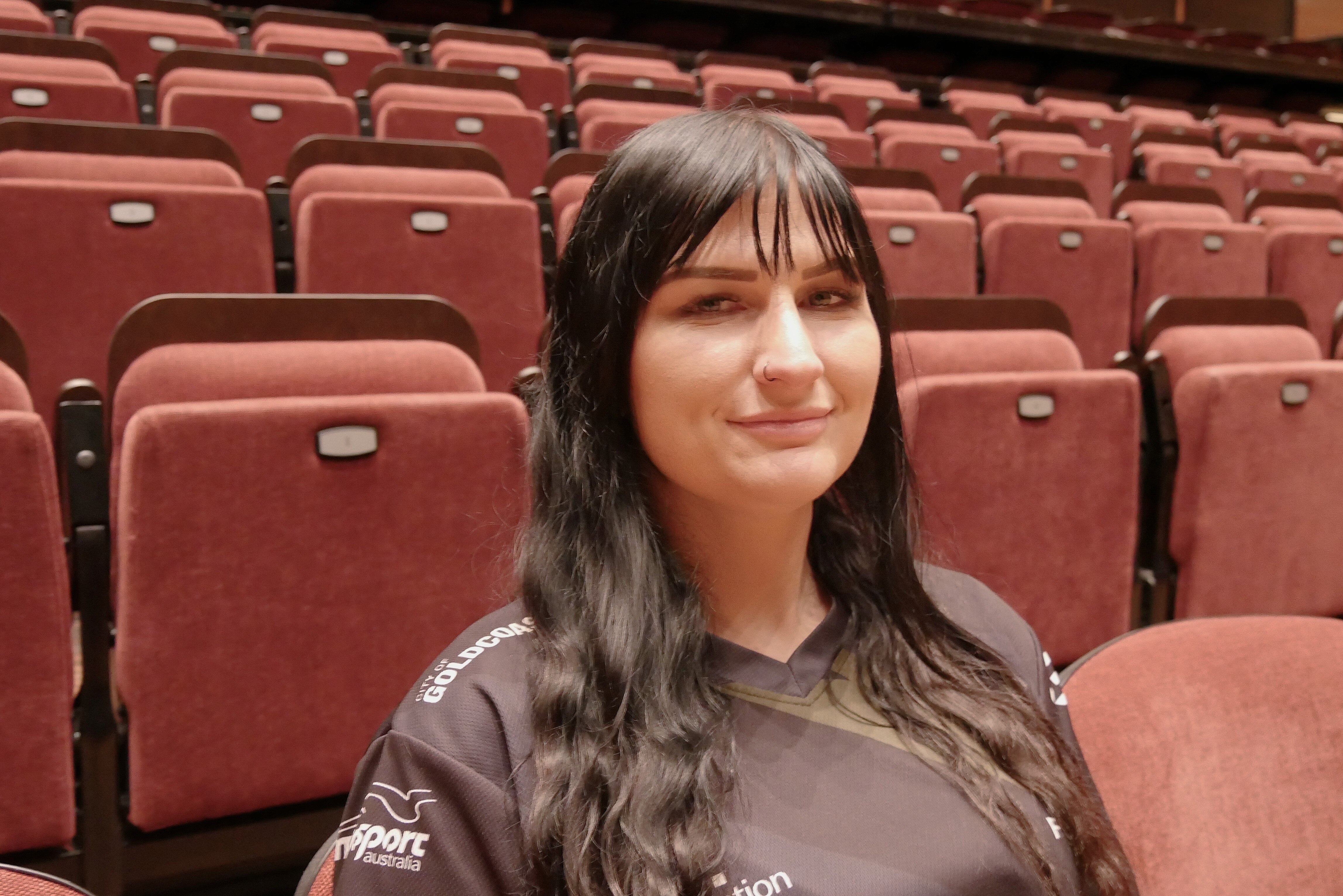 Woman with black hair and a black shirt sitting on a red chair with rows of red chairs behind her