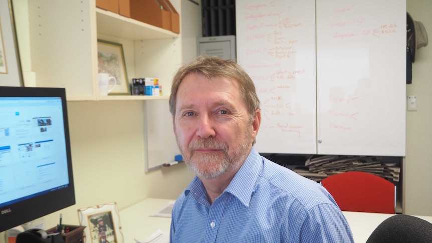 A university professor sitting in front of a computer with a whiteboard in the background