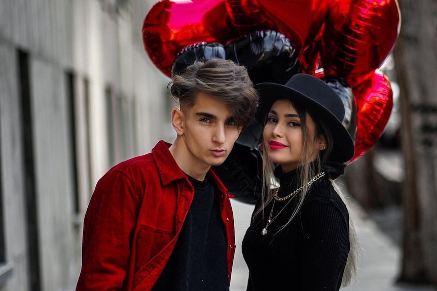 A young, well-dressed good-looking Iranian couple pose on a street with red and black heart-shaped balloons.
