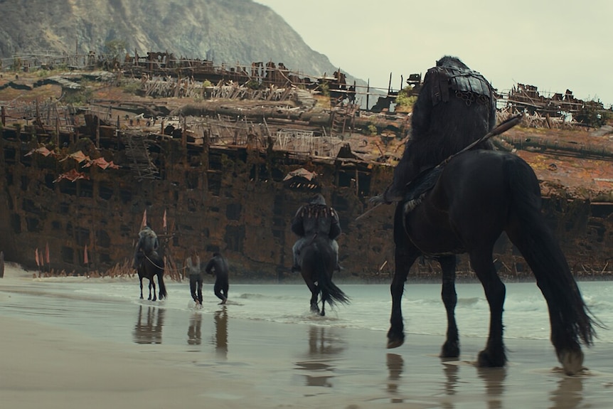 Apes ride horses toward a giant shipwreck on a beach. 