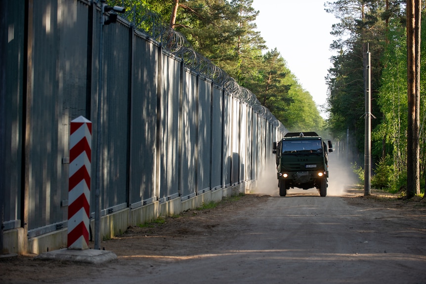A military truck drives along a tall, steel fence in the middle of a forest. 