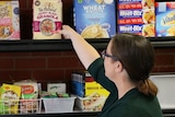 A woman reaches for a packet of cereal on a shelf in a store.