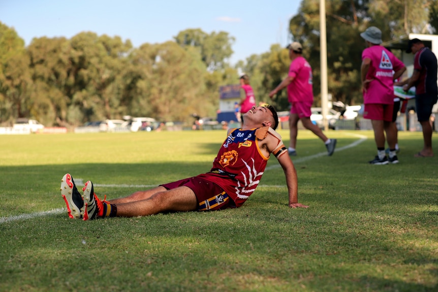 Young footballer stretches on sidelines of green grass football oval, wincing in pain. 