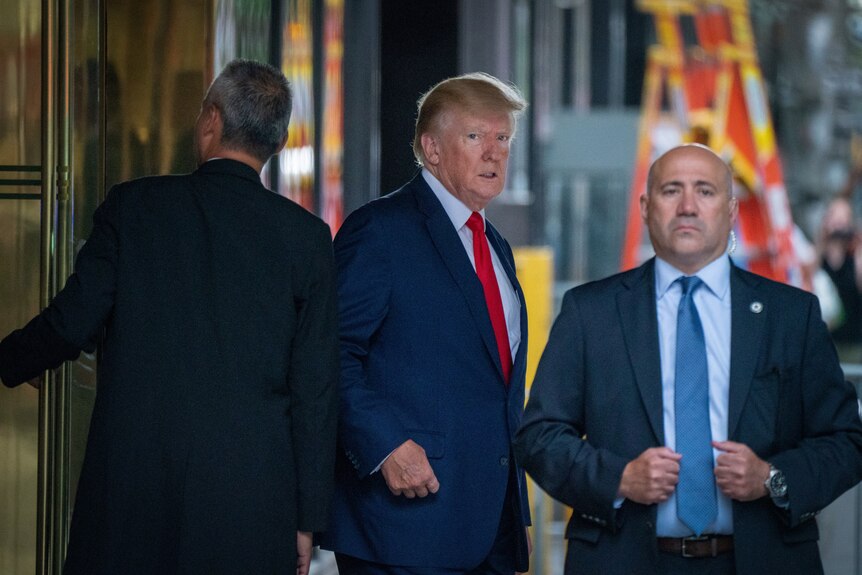 L'ancien président Donald Trump a l'air stressé dans un costume bleu et une cravate rouge avec deux hommes en costume de chaque côté de lui